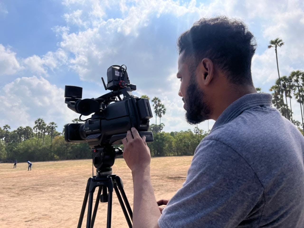 Camera operator filming a cricket match under a blue sky